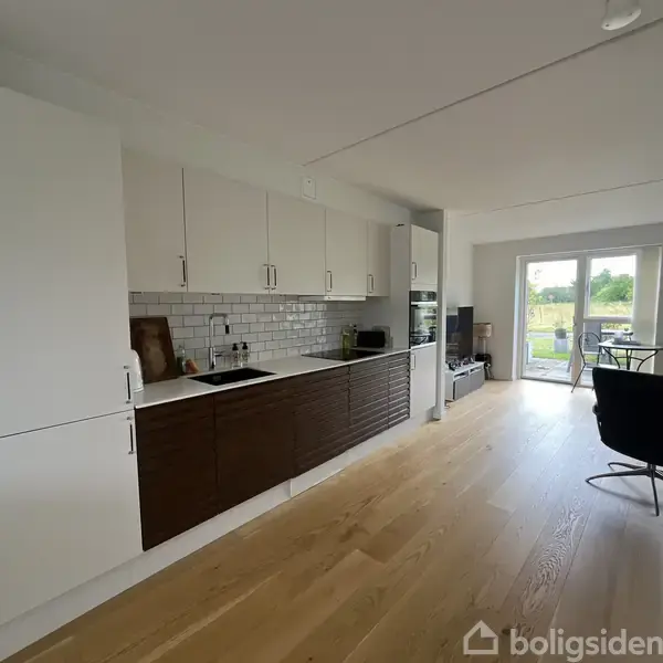 Køkken med hvide og mørke skabe placeret mod væggen, flankeret af et spiseområde med en glasdør, der åbner ud til haven. Bræddegulv og minimalistisk indretning.