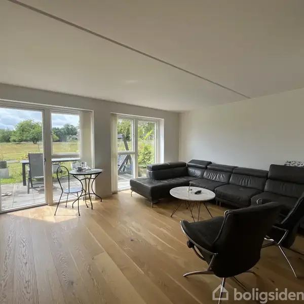Sort lædersofa står i en lys stue med trægulv. Store vinduer åbner ud mod en terrasse med udsigt over en grøn have. To sorte stole og et lille rundt bord.