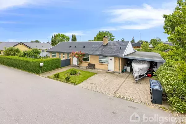 En étplans murstenshus med sort tag har en overdækket carport med en dækket båd. En forhave med græs og roser omgiver huset. Det ligger langs en stille villavej.