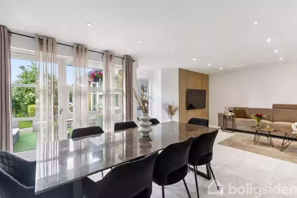 Spisebord står dækket med sorte stole i en moderne stue. Billedet har også en sofa, tv på væggen og store vinduer, der viser en have udenfor.