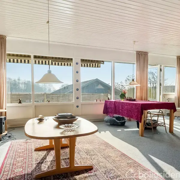 Et lyst, rummeligt rum med spisebord i midten og et bord dækket med rød dug tæt på vinduerne med udsigt til en have. Sollys strømmer ind gennem store vinduer.
