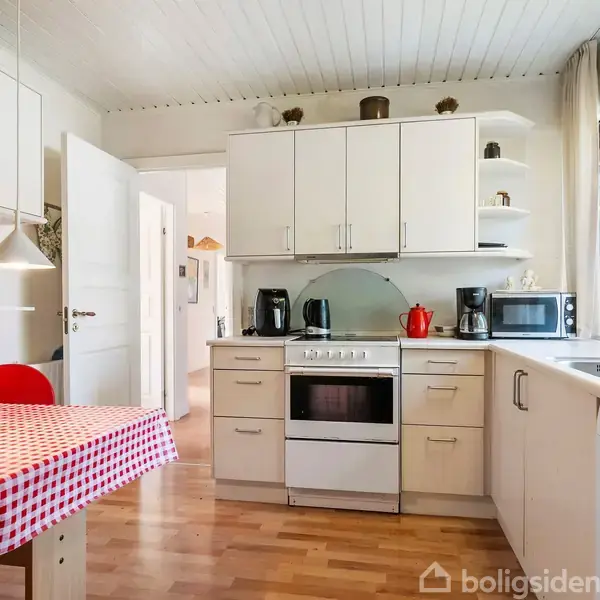 Køkken med hvidt interiør og trælookgulv. Bord med rød-og-hvidternet dug står foran skabe. Elkedel og mikrobølgeovn på bordpladen. Stor vindue giver udsigt til grøn beplantning.