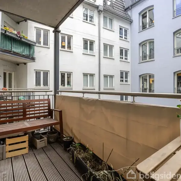 En balkon med træbænk og plantekasser, stående mod en beige afskærmning. I baggrunden ses en indre gård omgivet af hvide murstensbygninger. Tekst på krukke: "I'LL BE WITH YOU THIS DAY & ALWAYS".