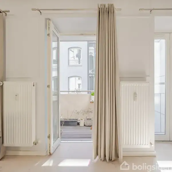 Dør står åben ind til en lys altan med planter, omgivet af hvide gardiner og radiatorer. Lyst rum med en del af en sofa synlig til venstre.