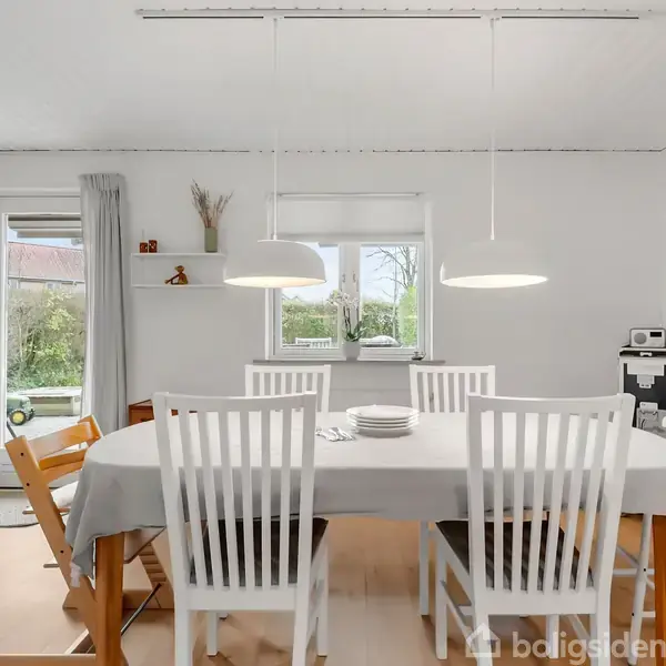 Et spisebord med hvid dug er omgivet af seks hvide stole. Bordet står i et lyst rum med træmøbler, to hængelamper over bordet og en stor have udenfor vinduet.