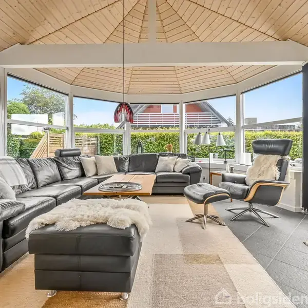 En stor, sort hjørnesofa er centreret i et lyst rum med store vinduer, der giver udsigt til en terrasse og have. En moderne lænestol og bord står foran sofaen.