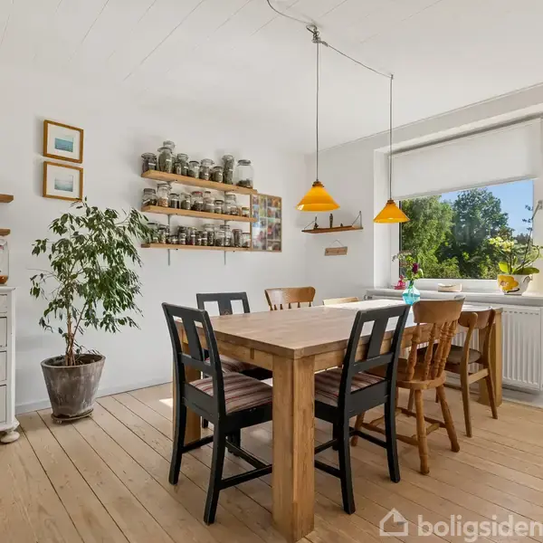 Træbord med seks stole står i en lys spisestue. To orange lamper hænger over bordet. I baggrunden ses en stor plante, hylder med glas og et stort vindue.