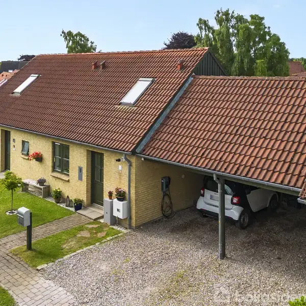Et rækkehus med rødt tegltag og gule murstensvægge står ved en grusbelagt indkørsel. En bil er parkeret i carporten. Græsplæne og grønne buske omkranser huset.