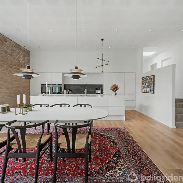 Rundt spisebord med sorte stole står på et rødt orientalsk tæppe. Moderne køkken i baggrunden. Rummets lyse vægge har kunstværker, og der er synlige trappetrin til højre.
