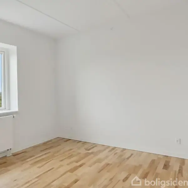 Et lyst værelse med trægulv og hvidmalede vægge, indeholder et vindue med udsigt til en grøn natur udenfor. Radiator er placeret under vinduet.