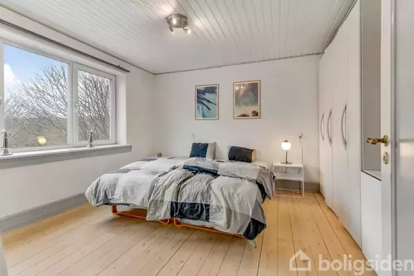 En seng står midt i et lyst soveværelse med trægulve. To malerier hænger på væggen over sengen. Et natbord med lampe står ved siden af sengen.