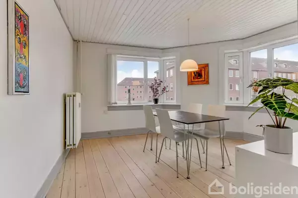 Et spisebord med fire stole står i et lyst rum med trægulve. Lampe hænger over bordet. Vinduer viser byudsigt. Vægmalerier og planter dekorerer rummet.