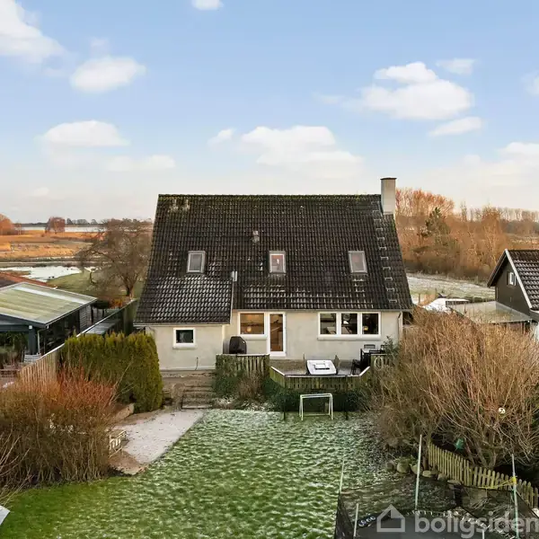 Et to-etagers hus står stille i en forstadshave omgivet af snoede buske og græsplæne, med naboer tæt ved og åbne marker i baggrunden under en blå himmel.