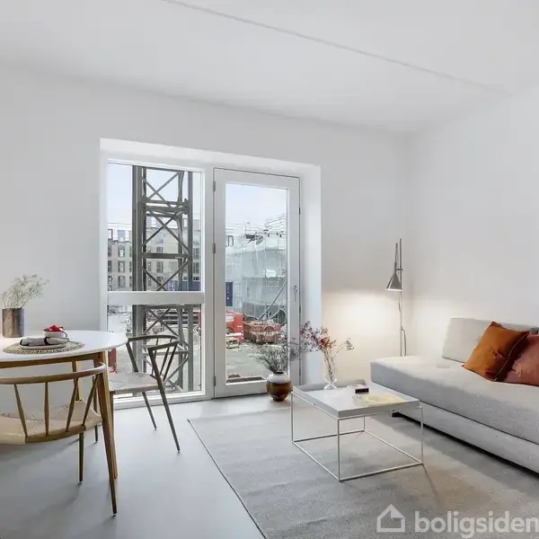 Stue med grå sofa og farvede puder ved siden af glasbord, står ved skydedør til altan. Rundt spisebord med dekorativ lampe og stole. Lys indretning.