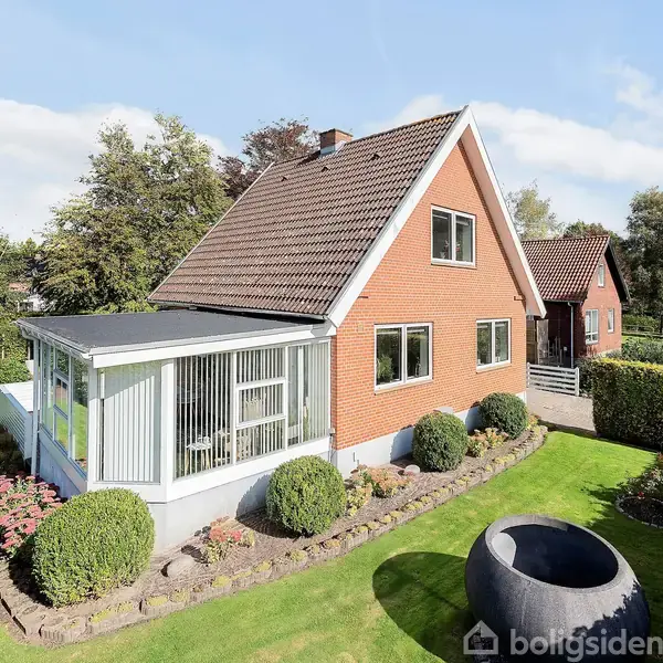 Et rødt murstenshus med gavl og vinduesparti. Det har en velplejet have med grønne buske og en stor græsplæne, omgivet af træer og buske.