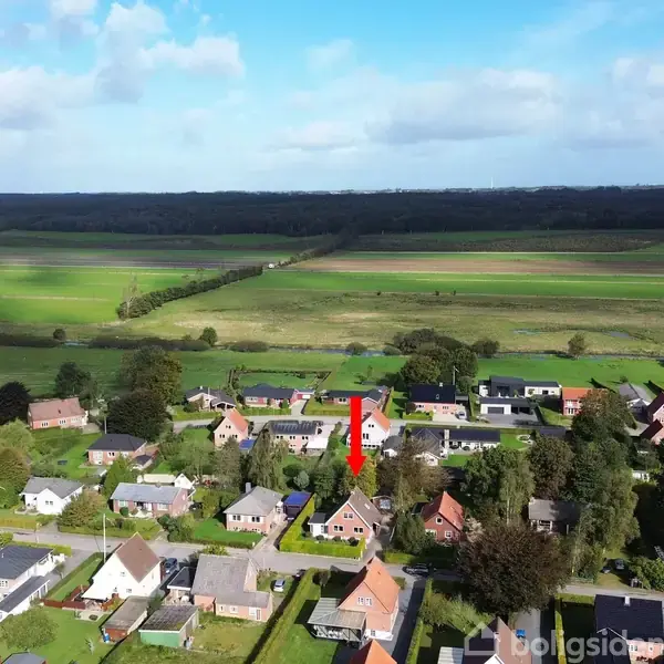 Et række huse ligger i en landsby med marker i baggrunden. En rød pil peger mod et specifikt hus blandt de mange omkringliggende bygninger og træer.