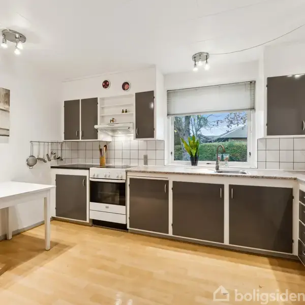 Et køkken med grå skabe og lyse træbordplader. Et hvidt bord står til venstre. Der er et stort vindue med udsigt, en ovn, og en opvaskemaskine.