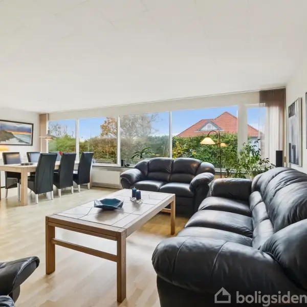 Sort lædersofaarrangement står i en lys stue med store vinduer, der giver udsigt til en have. Et spisebord med stole står i baggrunden, omringet af kunst på væggene.