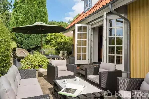 Terrassemøbler står stille på en solrig terrasse foran et hus med åbne dobbeltdøre. En stor grøn parasol og frodige buske omgiver området, skaber en hyggelig udendørs stemning.