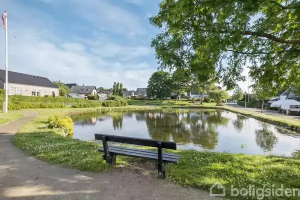 En bænk står foran en lille dam i et grønt parkområde omgivet af træer og huse. En flagstang ses til venstre, og himlen er blå med skyer.