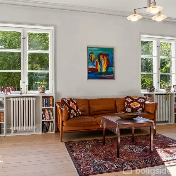 Stue med brun lædersofa står mod en hvid væg prydet med malerier. To vinduer giver udsigt til grønt løv. Rød tæppe under træbord. Reoler flankerer radiatoren.