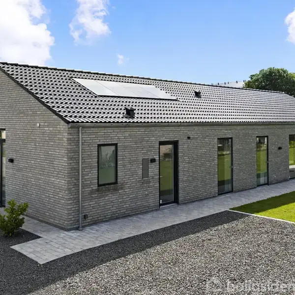 En moderne etplans murstensvilla med solpaneler på taget. Huset står på en anlagt grund med græsplæne og grusbelagt indkørsel, omkranset af blå himmel.