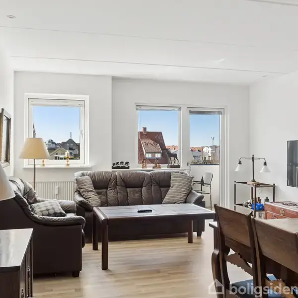 Stue indeholdende en mørk sofa og sofabord, lamper og vægkunst. Vinduer med udsigt over bygninger. Fladskærms-tv monteret på væggen. Spisebord med stole til højre.