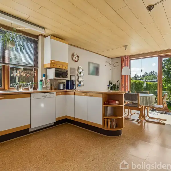 Et rummeligt køkken med hvide skabe og en stor dobbeltvask. Vinduet viser en have udenfor, mens et spisebord med stole står ved en glasdør til terrassen.