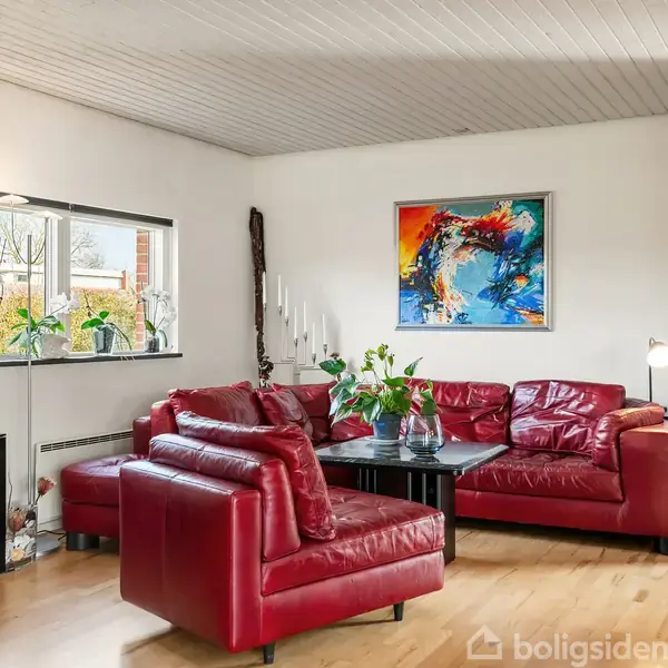 Rød lædersofa står i en stue, omgivet af et maleri på væggen med abstrakt kunst. Store planter og lysindfald fra et vindue skaber en lys og indbydende atmosfære.