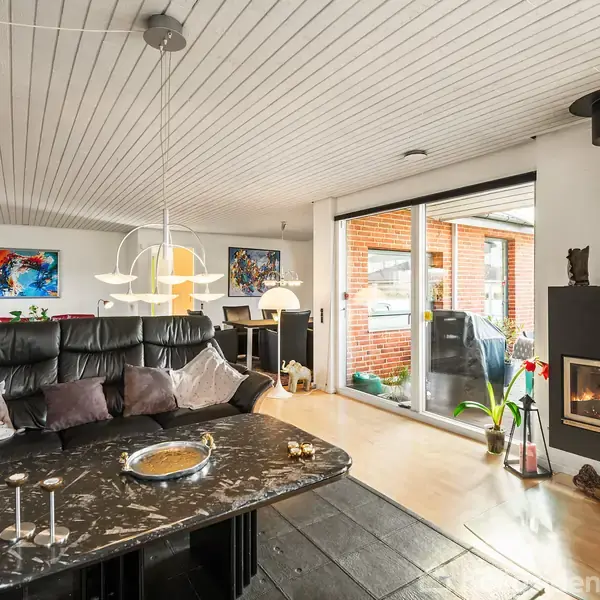 En sort lædersofa med puder står foran et sofabord med stearinlys, ved siden af en brændeovn i et moderne stueindretning med hvide lofter og store vinduer.