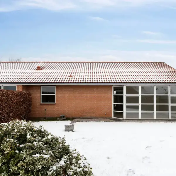 Et rødt murstenshus med et fladt tag står omkranset af sne. Store vinduespartier ses til højre. Buske dækket af sne omkranser huset, mens himlen er lys og let overskyet.