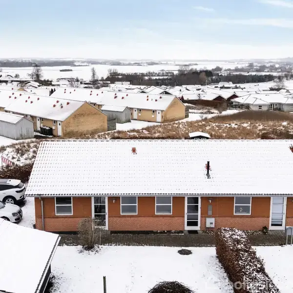 En rækkehusbygning med snedækket tag står i et vinterlandskab, omringet af flere lignende bygninger og sparsomme buske dækket af sne. Biler er parkeret ved siden af bygningen.