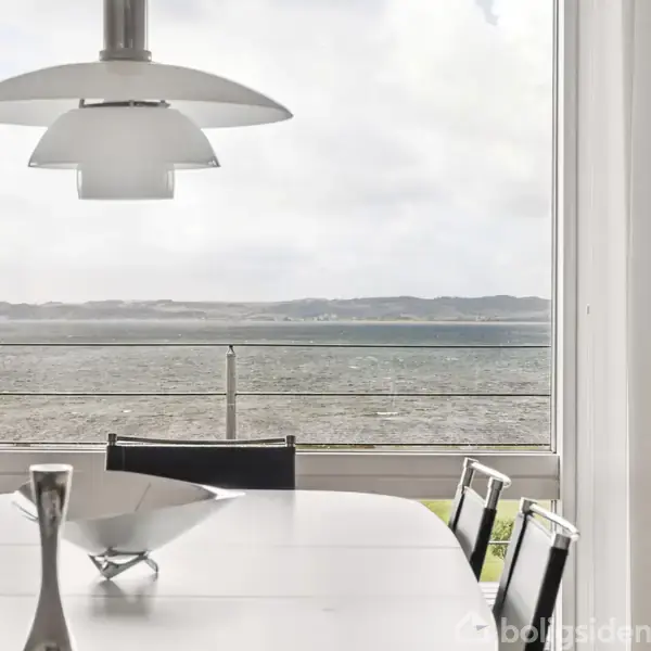 Et spisebord står foran et stort vindue med panoramaudsigt over en sø eller hav. En moderne lampe hænger over bordet. Udenfor ses et gelænder og bakkede landskaber i baggrunden.