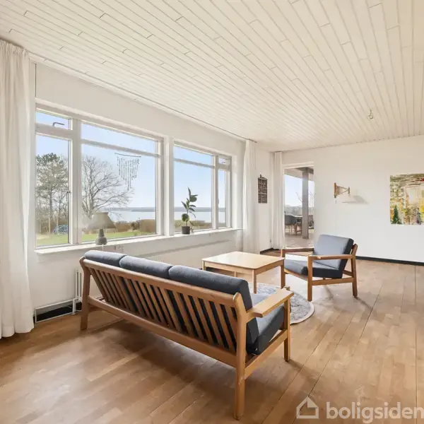 En stue med træmøbler står med en sofa og to lænestole omkring et sofabord. Store vinduer giver udsigt til grønne træer udenfor. En malet vægdekoration hænger på væggen.