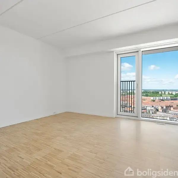 Et tomt rum har et trægulv og hvide vægge. En stor døråbning med glas fører ud til en balkon med udsigt over en by og en blå himmel.
