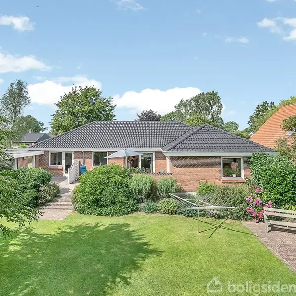 Et enfamiliehus i en havekontekst med blomsterbede og en græsplæne. Huset har røde mursten, sort tag og et flisebelagt område. Buske og træer omgiver ejendommen.