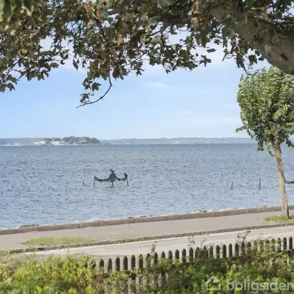 En fredelig sø omgivet af træer og en sti i forgrunden. Vandet strækker sig mod en ø i horisonten. Der står et hegn langs stien.
