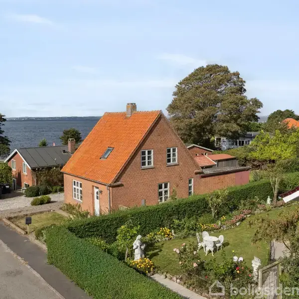 Et rødt murstenshus med orange tag står i en velplejet have med buske og blomster, placeret i et kystnært område med havudsigt.