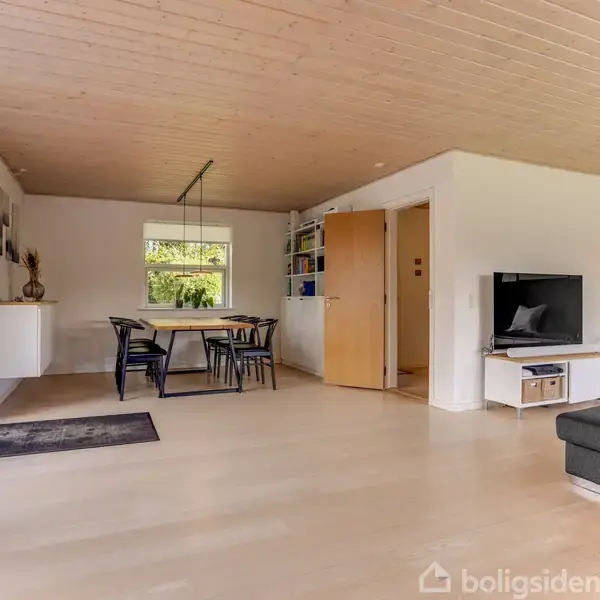 Stue med gråt sofaarrangement mod højre hjørne, TV på væggen, spisebord med sorte stole ved et vindue til venstre, lyst trægulv og træloft, vinduer mod haven.