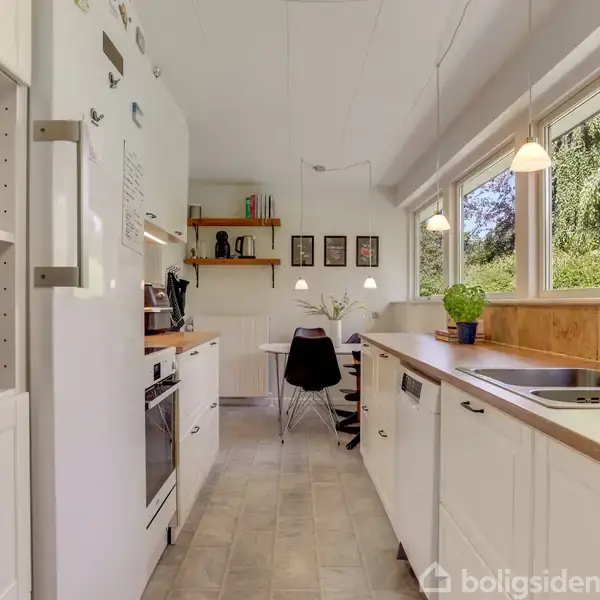 Køkken med hvide skabe, hvor en opvaskemaskine og ovn er integreret. Rummet er lyst med store vinduer, planter og spiseplads i baggrunden. Hylder med bøger og dekorative elementer.