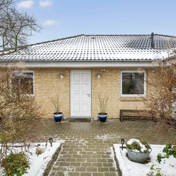 Et hus med gulstensfacade står stille omgivet af sneklædt have og flisebelagt sti. To vinduer og en hvid dør er synlige. Plantekrukker placeret ved indgangen.