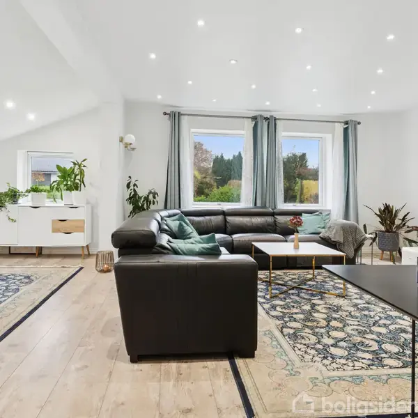 Sort hjørnesofa står i et lyst, rummeligt stueområde omgivet af tæpper og planter. Store vinduer med gardiner tilføjer naturligt lys. Et TV og diverse småborde flankerer sofaen.