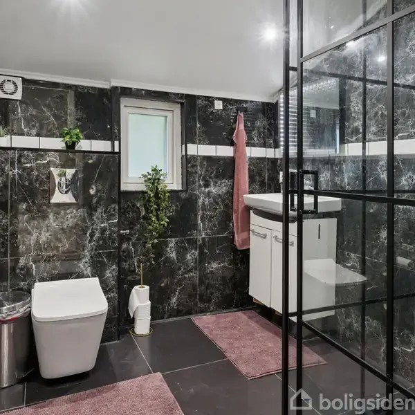 Toilet og håndvask er placeret i et badeværelse med sorte marmorerede fliser. Der er et lille vindue, to lyserøde måtter og et håndklæde, samt en affaldsspand og toiletpapirholder.