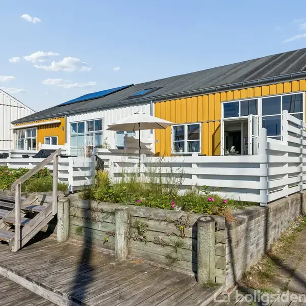 En gul bygning med store vinduer har en forhave med hvidt hegn og beplantning. Træterrassen foran fører til indgangen. Sollys skinner på strukturen, omgivet af andre huse.