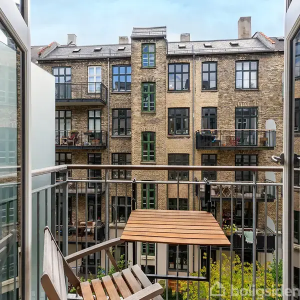 Balkon med træbord står stille foran en baggård med murstensbygninger og flere altaner, set fra en højere etage i en byejendom.
