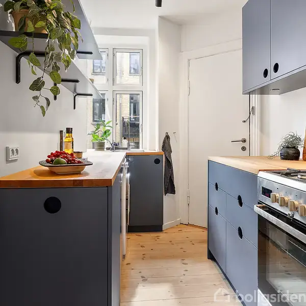 Et moderne køkken med blå skabe og træbordplader. En komfur står til højre, omgivet af køkkenredskaber. En plante og andre dekorationer pryder hylderne. Lyset strømmer ind fra vinduerne.