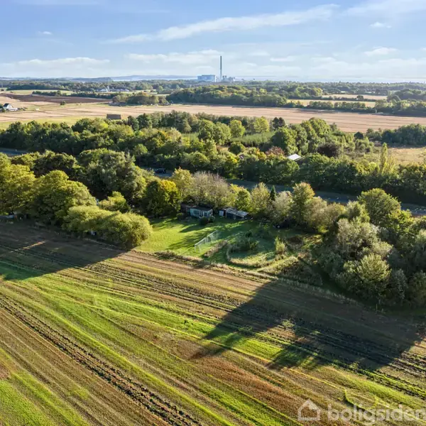 Landskab med marker strækker sig ud under en klar himmel. Træer omkranser et område i midten, mens bygninger og en fabriksskorsten er synlige i det fjerne.