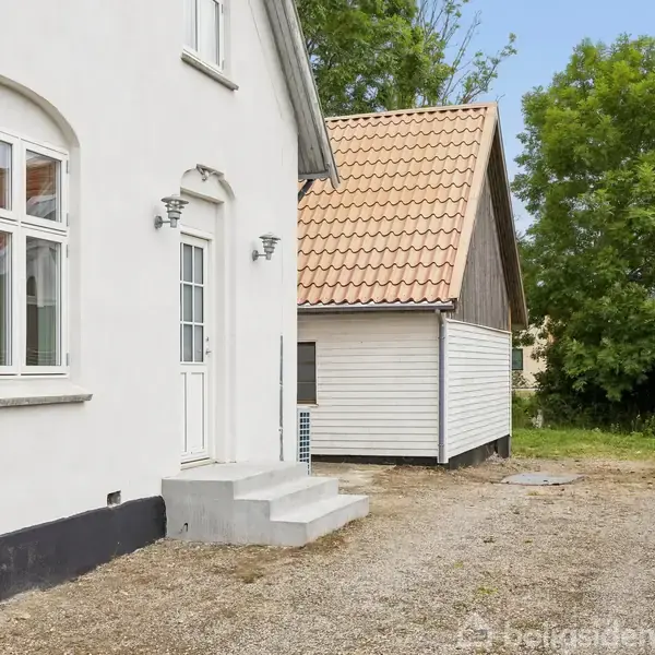 En hvid facadebygning med store vinduer og en indgangsdør med trappetrin fører ud til en gårdsplads. Ved siden af står en lille bygning med et rødt tegltag og træbeklædning.