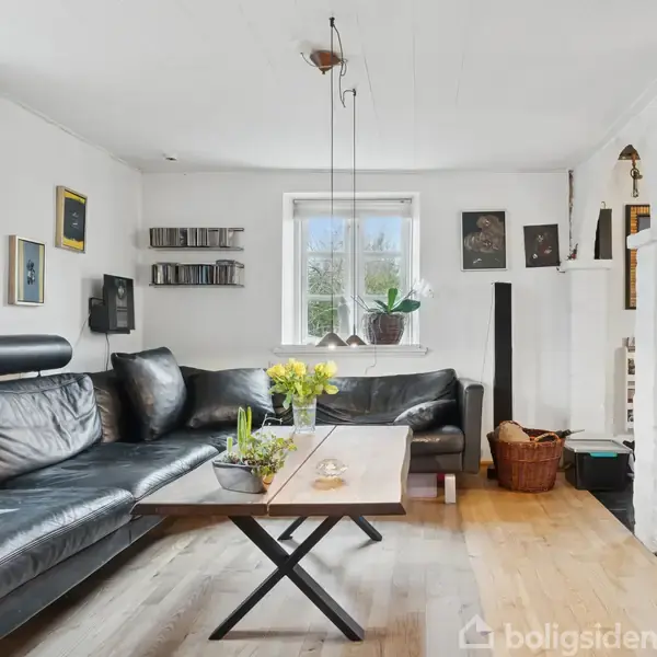 Sort lædersofa står i et hyggeligt stuehjørne med hvide vægge og træplankegulv. Et træbord foran sofaen har en blomsterdekoration. Billeder og hylder pryder væggene.