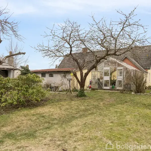Et gult hus med sort tag står omgivet af en have og et lavt hegn. Der ses andre huse i baggrunden, og himlen er blå.
